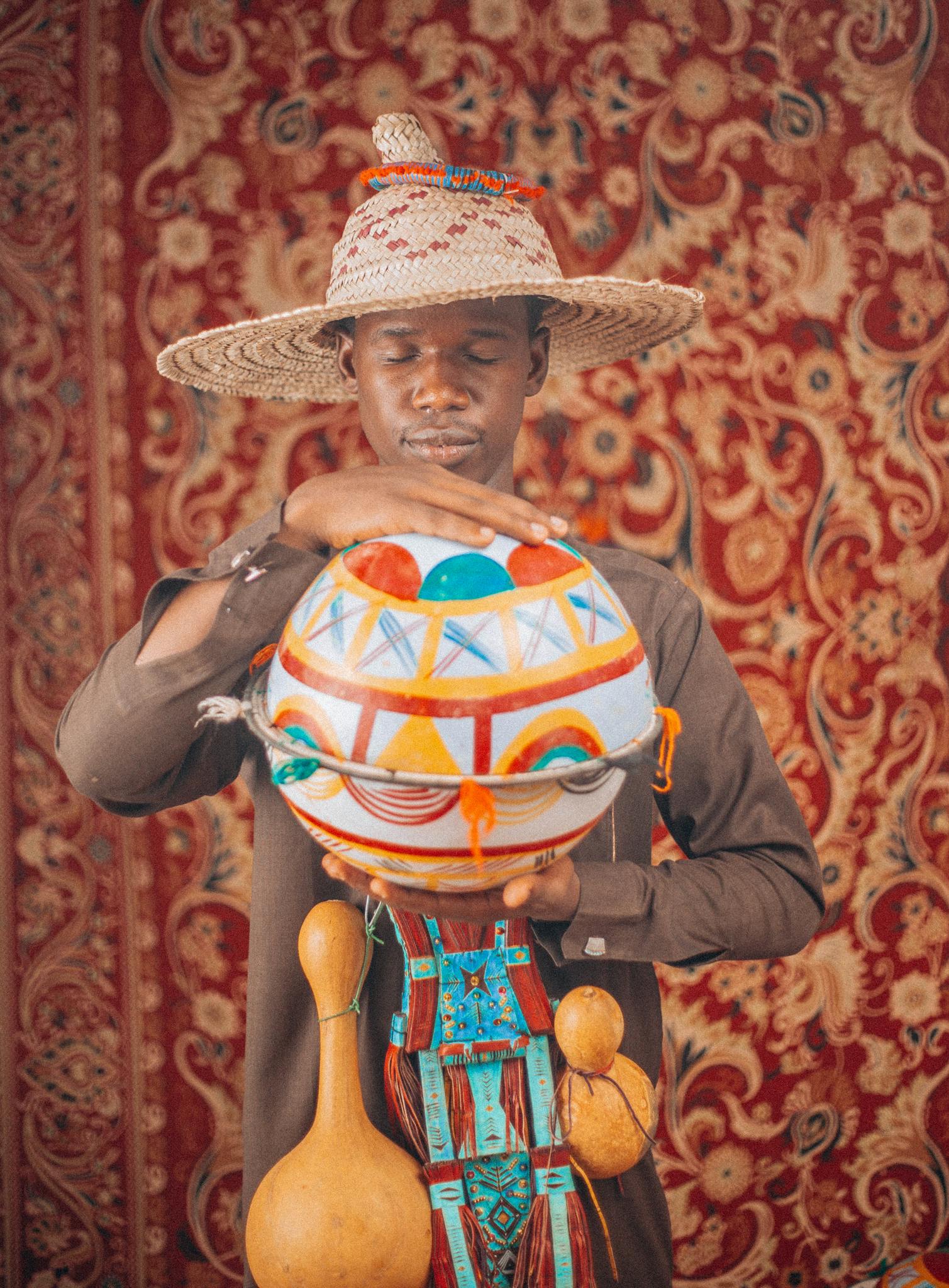 Man wearing a traditional hat holding an African decorated calabash in front of ornate backdrop.